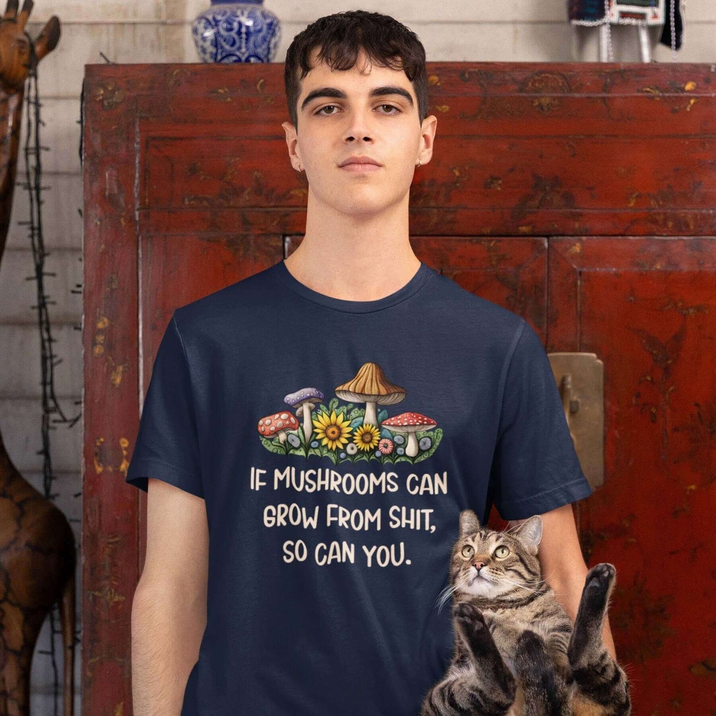 Man holding a cat while wearing a navy blue t-shirt. The shirt has images of mushrooms and the phrase If mushrooms can grow from shit so can you printed on the front.