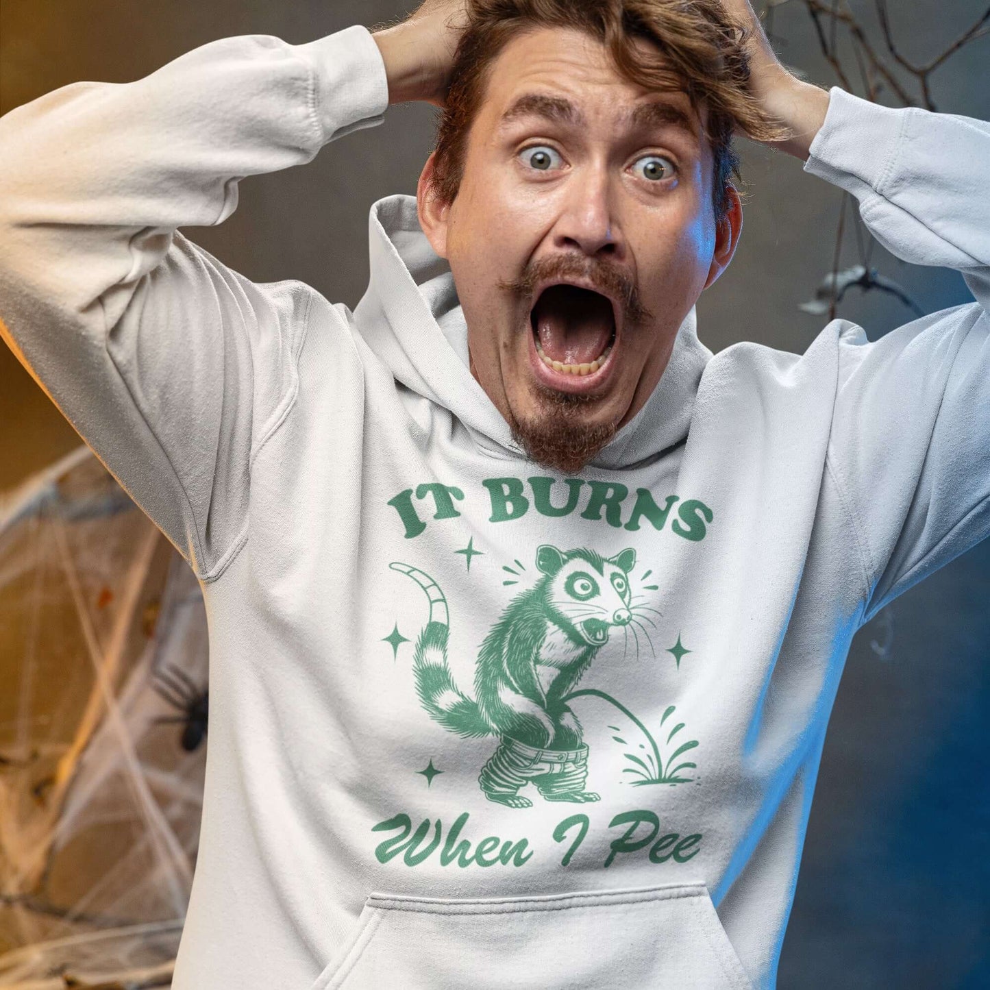 Man wearing a white hoodie sweatshirt that has graphic of an opossum peeing and the words it burns when I pee printed on the front.