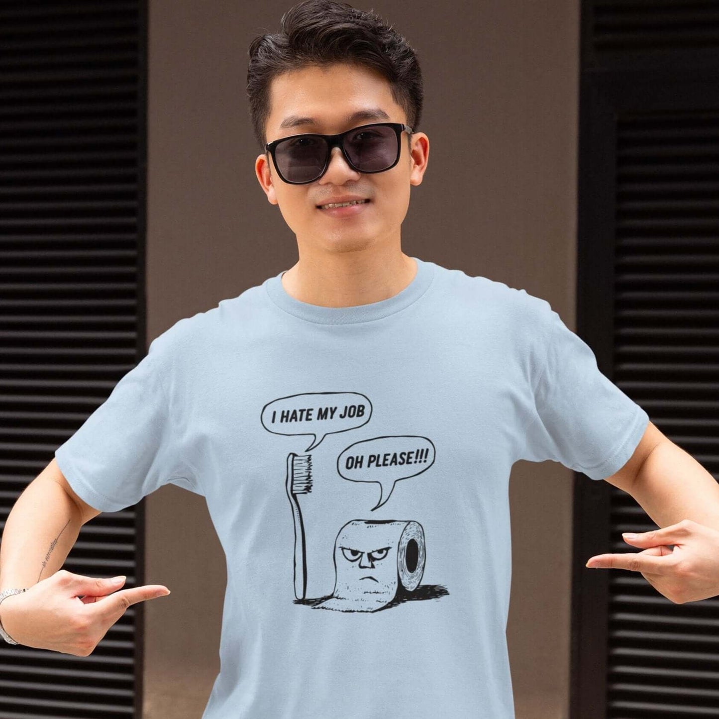 Man wearing a light blue t-shirt with a cartoon graphic on the front. The cartoon is a line drawing of a toothbrush and roll of toilet paper. Both have speech bubbles above . The toothbrush says I hate my job and the toilet paper says Oh please.