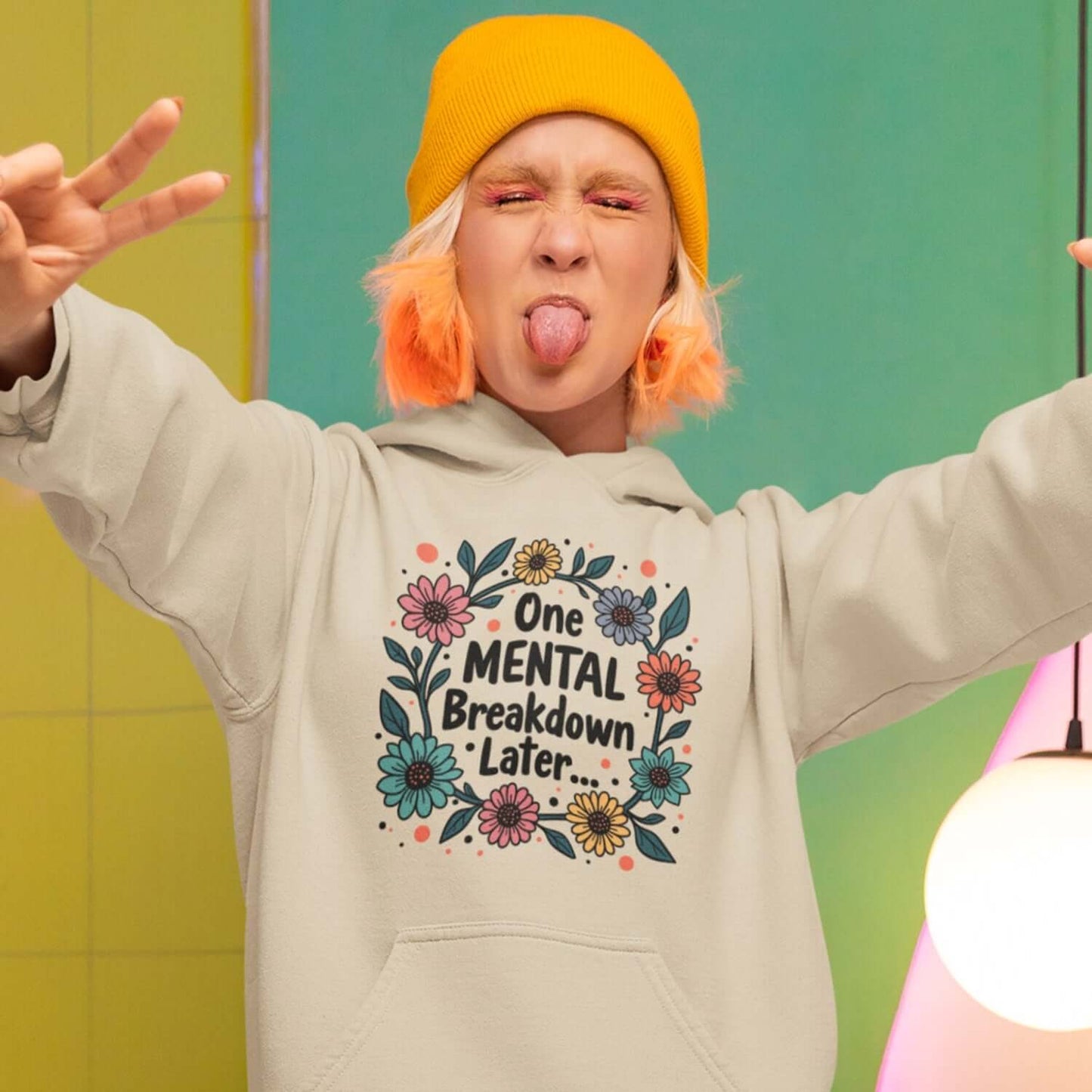 Woman making a funny face wearing a sand color hoodie sweatshirt with a multi color floral wreath design printed on the front. The words One mental breakdown later are printed in the center of the wreath.