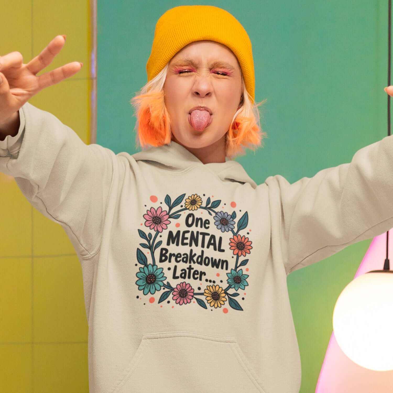 Woman making a funny face wearing a sand color hoodie sweatshirt with a multi color floral wreath design printed on the front. The words One mental breakdown later are printed in the center of the wreath.