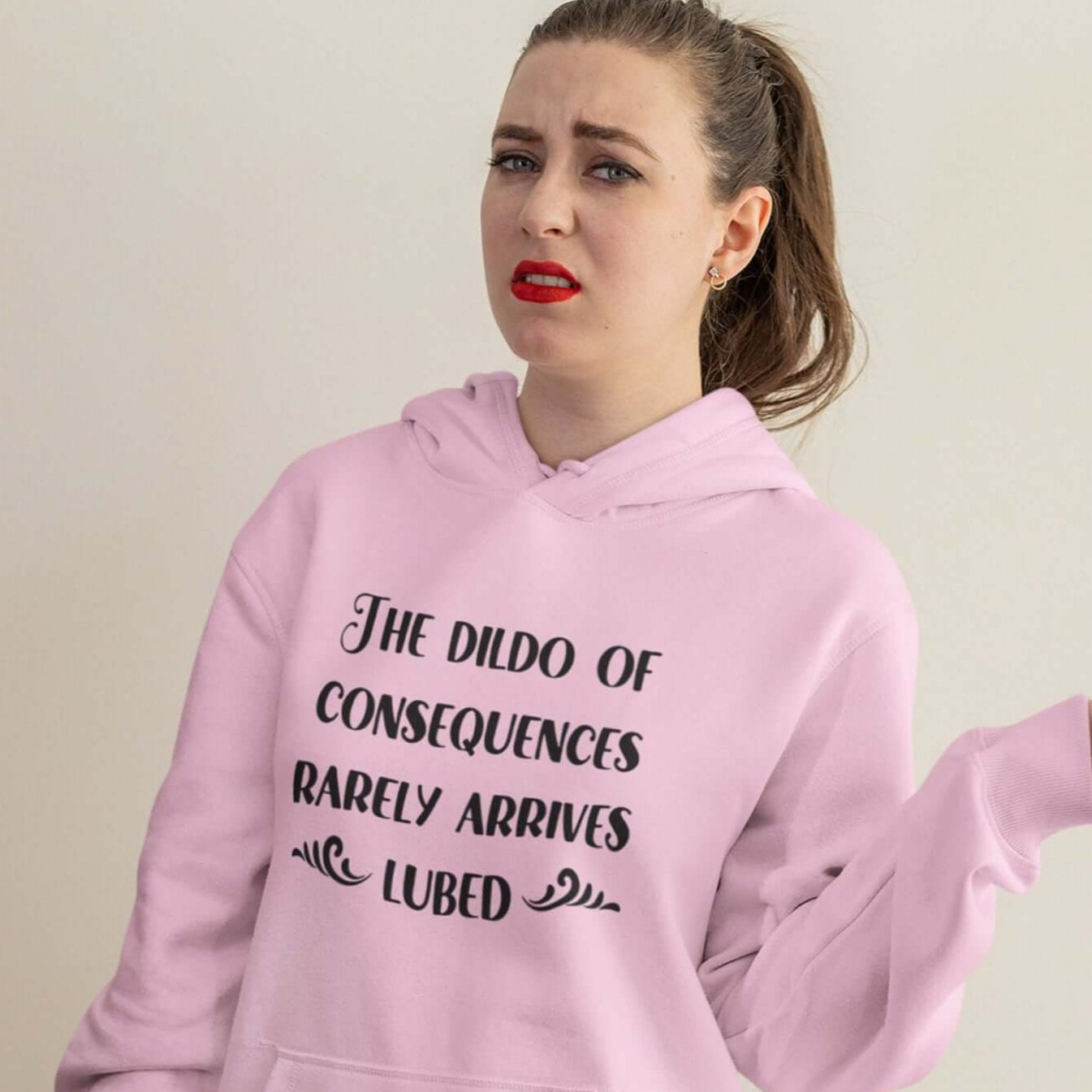 Woman wearing a light pink hoodie sweatshirt with the phrase The dildo of consequences rarely arrives lubed printed on the front.