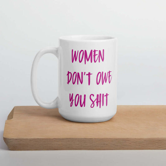 White ceramic coffee mug with the words Women don't owe you shit printed on both sides in pink.