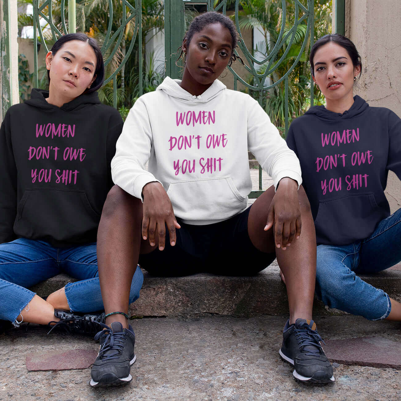 Three fierce looking women sitting next to each other. They are all wearing a hoodie sweatshirt with the words Women don't owe you shit printed on the front in pink.