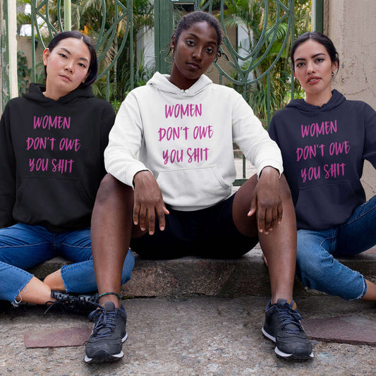 Three fierce looking women sitting next to each other. They are all wearing a hoodie sweatshirt with the words Women don't owe you shit printed on the front in pink.