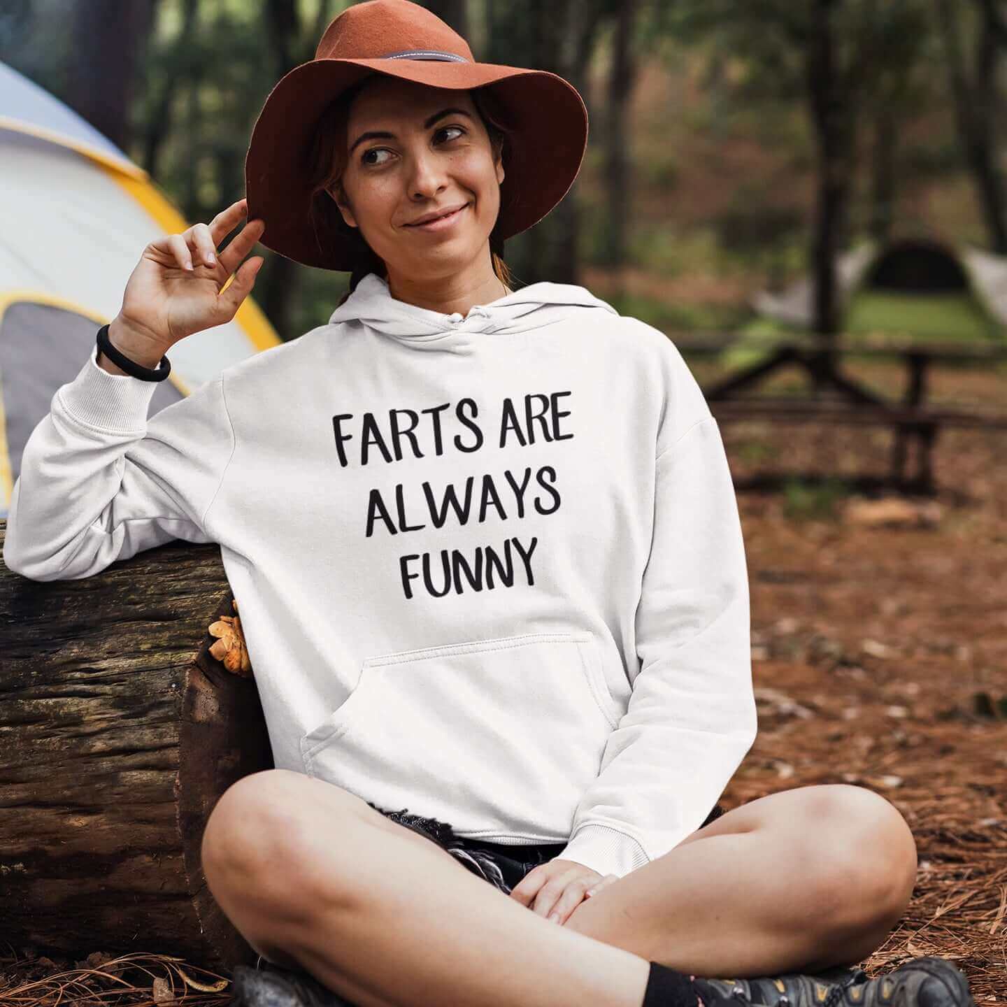 Woman in a camping setting wearing a white hoodie sweatshirt with the phrase Farts are always funny printed on the front.