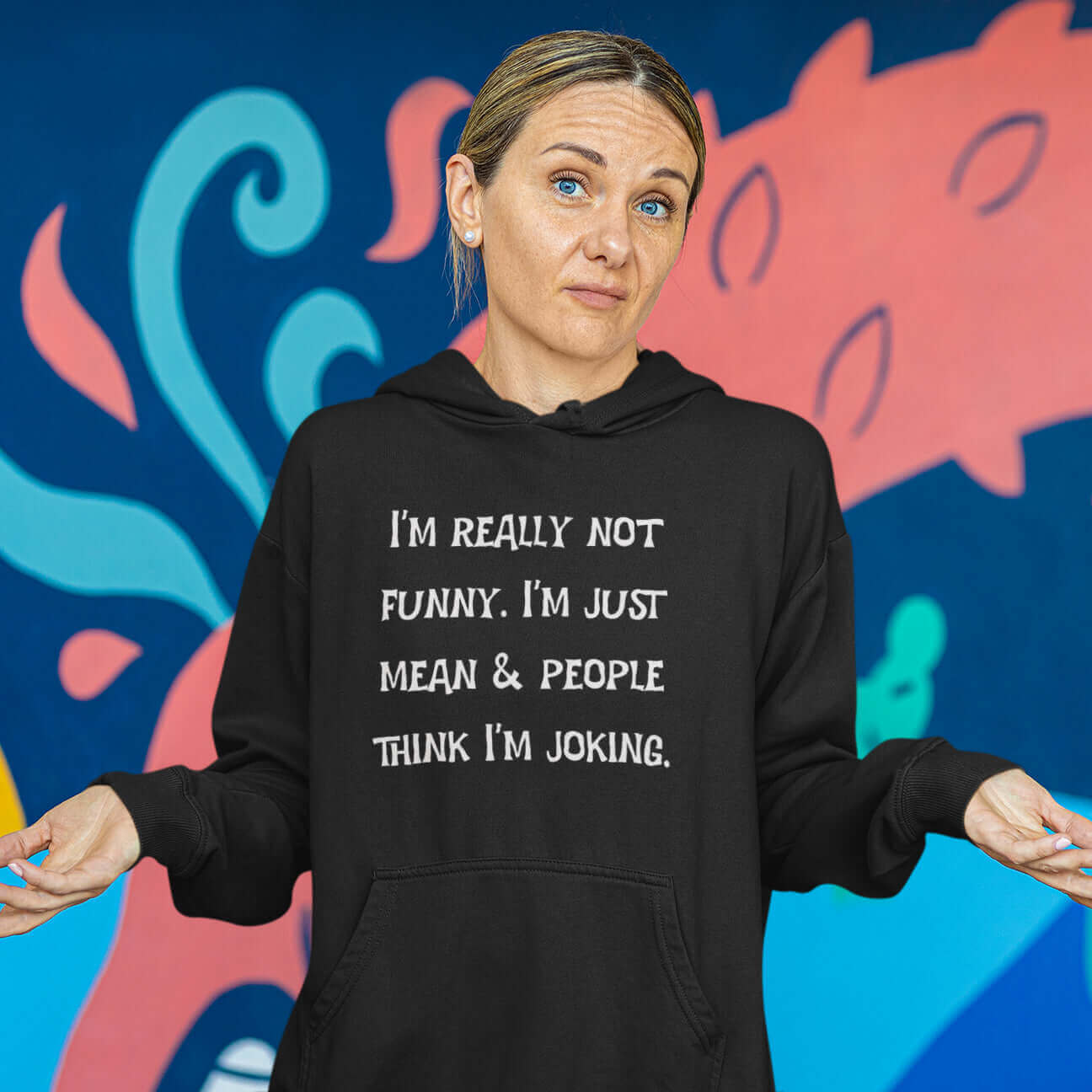 Shrugging woman wearing black hooded sweatshirt with the phrase I'm really not funny. I'm just mean & people think I'm joking printed on the front.