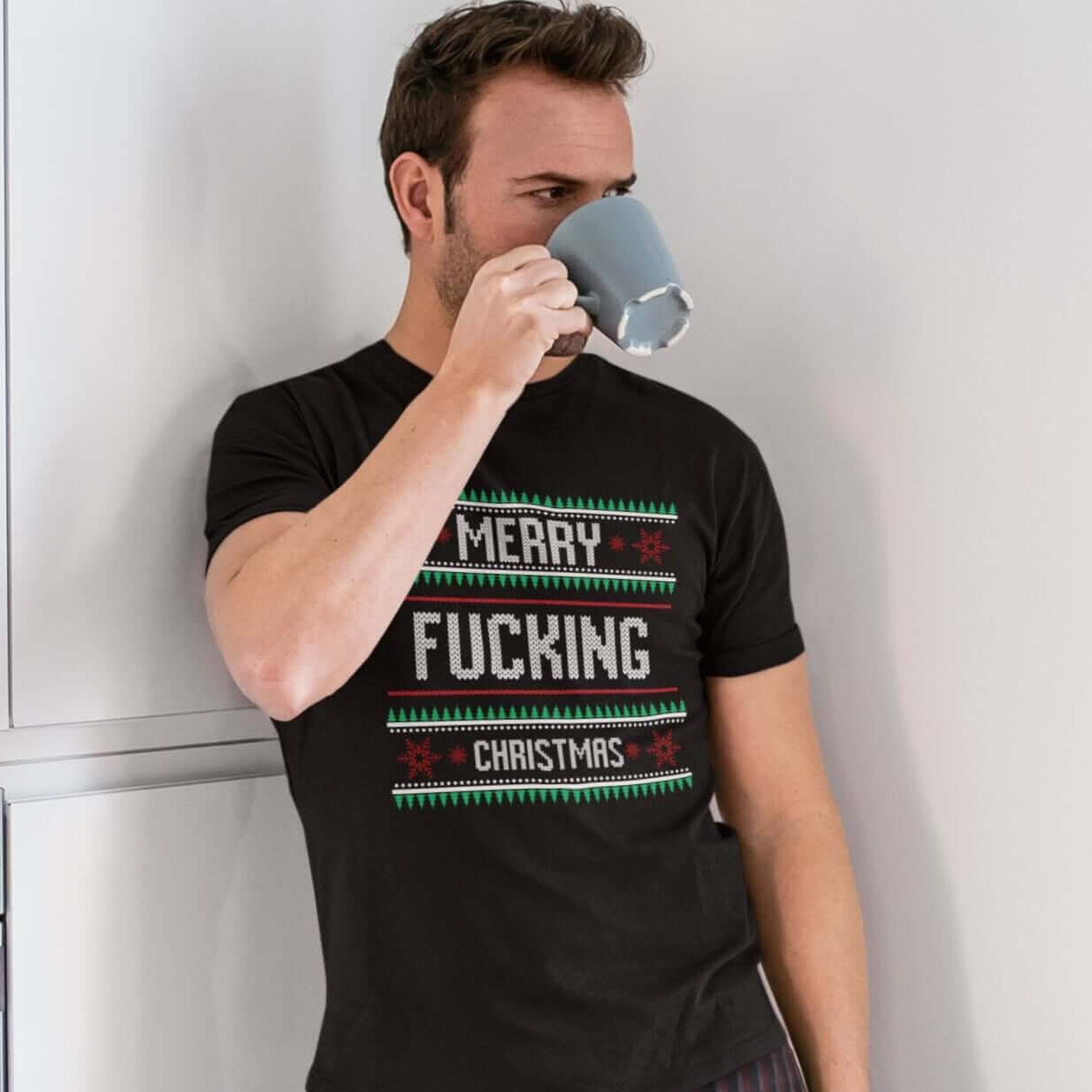 Man drinking coffee wearing pajama pants and black t-shirt with the phrase Merry fucking Christmas printed on the front in sweater print font.