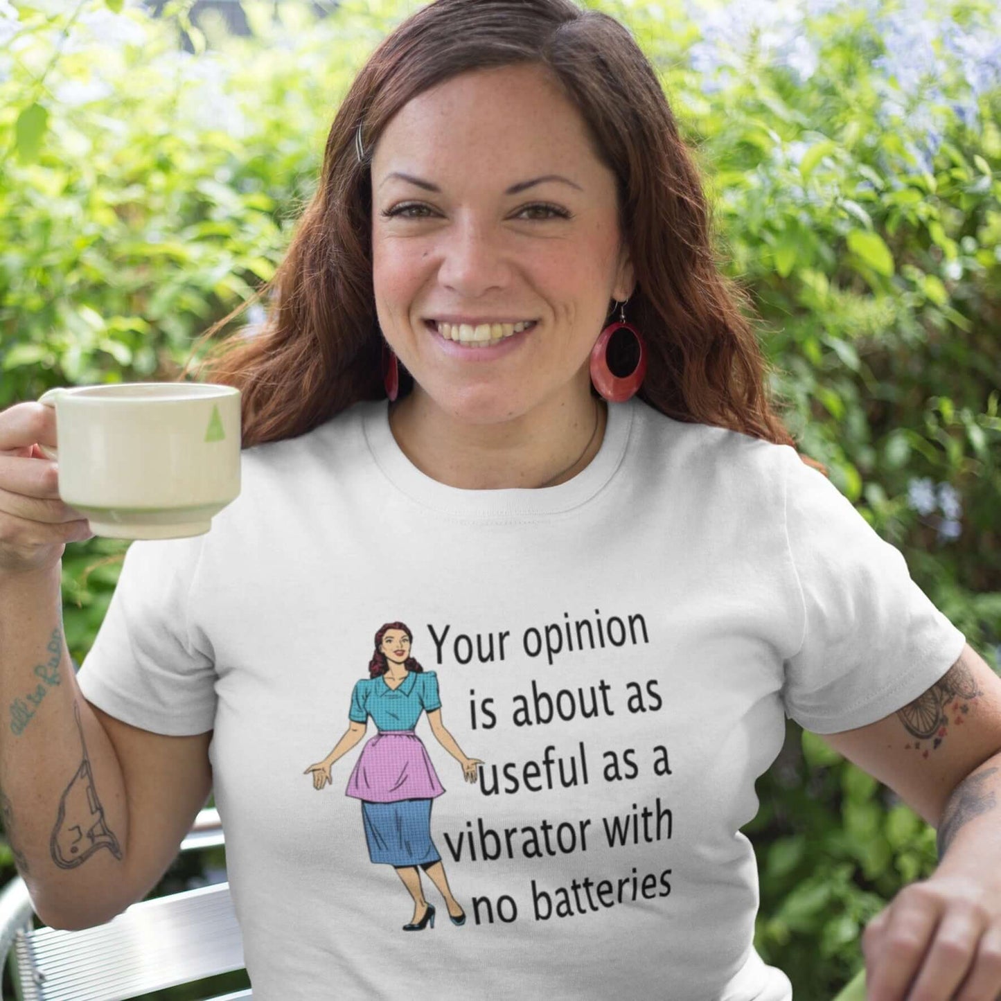 Woman sitting at a cafe drinking a cup of coffee. She is wearing a white t-shirt with an image of a retro woman and the phrase Your opinion is about as useful as a vibrator with no batteries printed on the front.