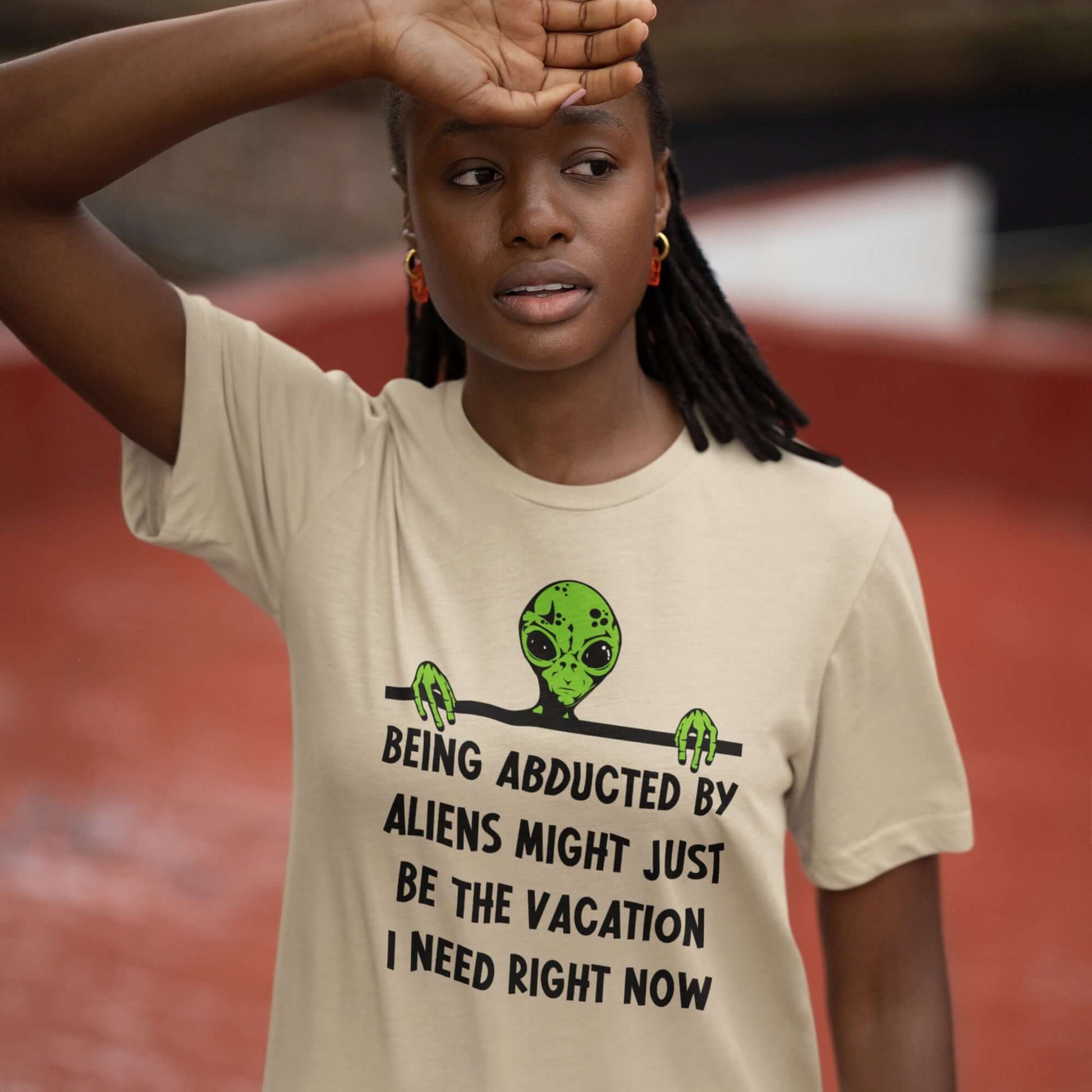 Woman wearing a sand color t-shirt with her hand on her forehead. The t-shirt has an image of a green alien and the phrase Being abducted by aliens might just be the vacation I need right now printed on the front of the shirt.