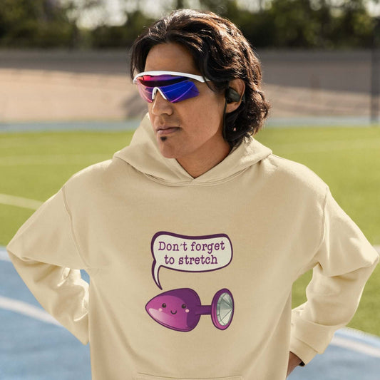 Man wearing a Sand color hoodie sweatshirt that has image of a purple anal plug with a smile face with a speech bubble above with the words Don't forget to stretch. The graphics are printed on the front of the hoodie.