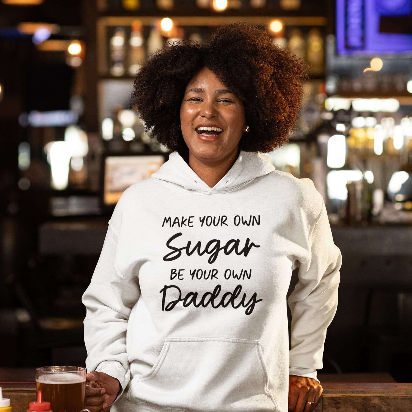 Smiling woman in a bar setting wearing a white hoodie sweatshirt. The sweatshirt has the phrase Make your own sugar Be your own Daddy printed on the front.