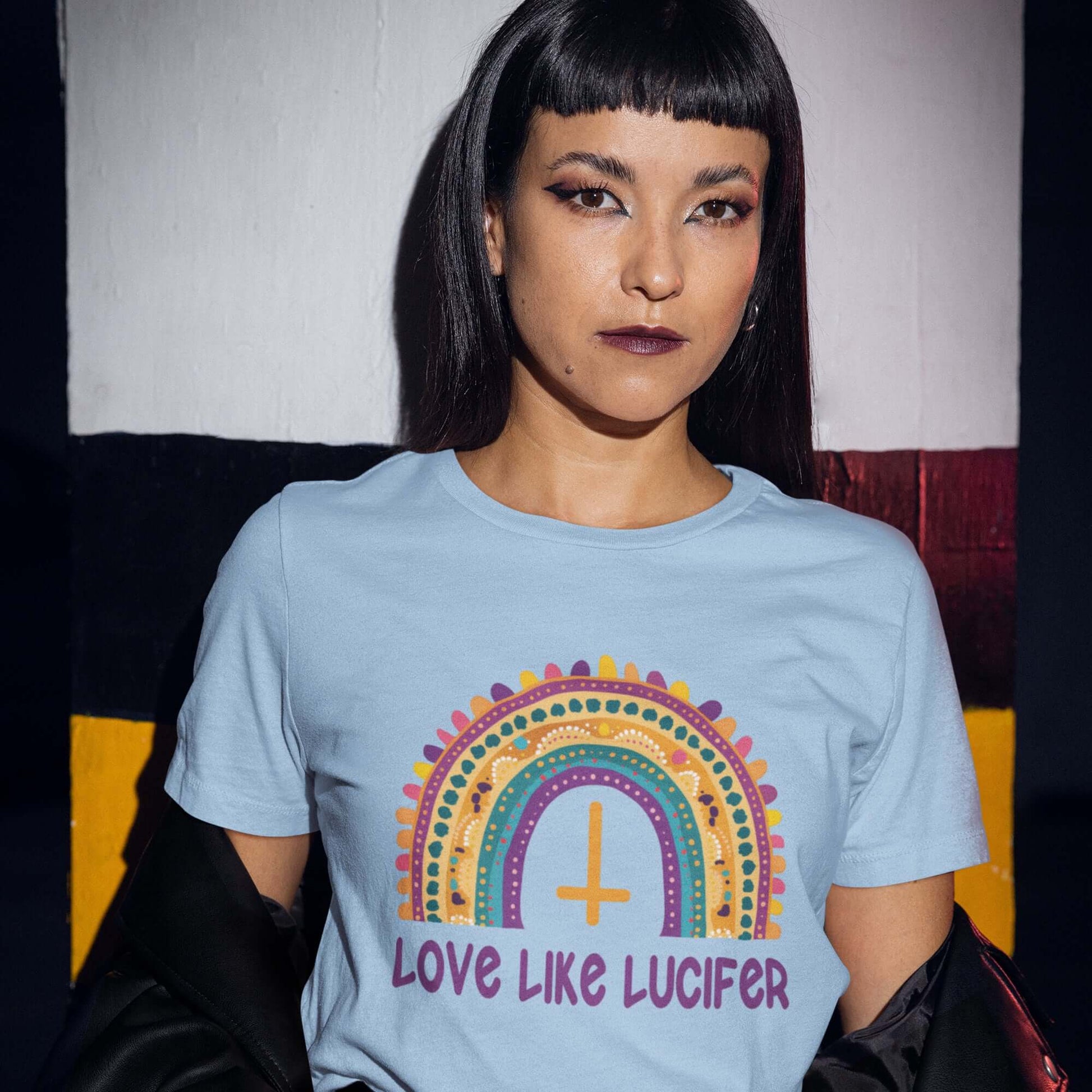 Woman wearing a light blue t-shirt with an image of a boho style rainbow. There is an upside down cross in the center arch of the rainbow and the phrase Love like Lucifer printed under. The graphics are printed on the front.