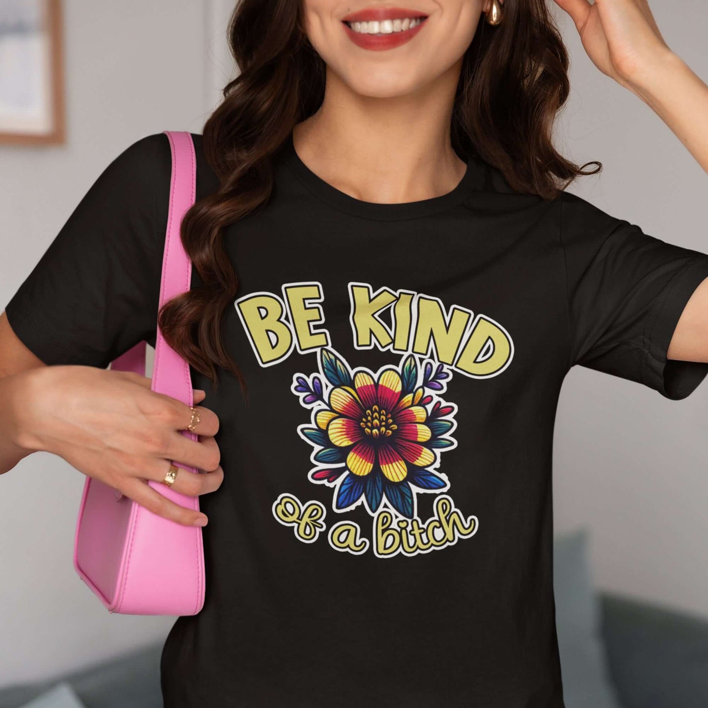 Woman wearing a black t-shirt with an image of a flower and the words Be kind above the flower in yellow bold block font. The words Of a bitch are smaller in script font under the flower. The design is printed on the front of the shirt.