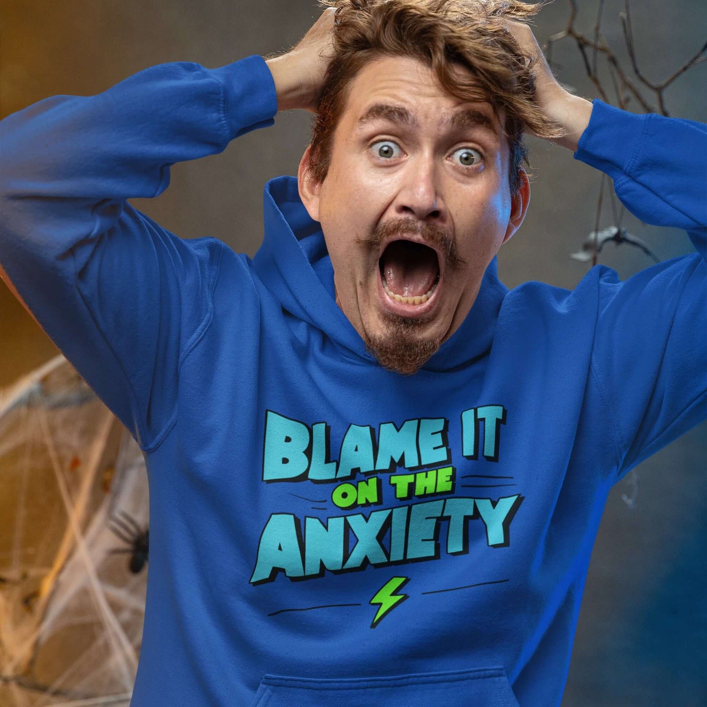 Freaked out man wearing a royal blue hoodie sweatshirt with the phrase Blame is on the anxiety printed on the front. The graphics are bold and in aqua and lime green.