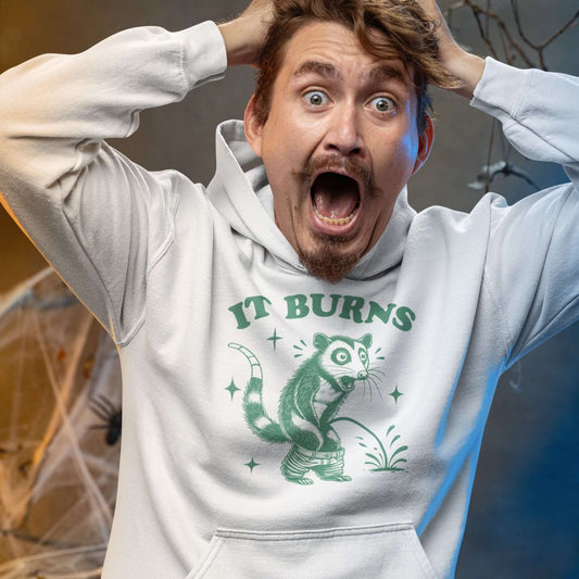 Man screaming while wearing a white hoodie sweatshirt with an image of an opossum peeing. The words It burns are printed above.