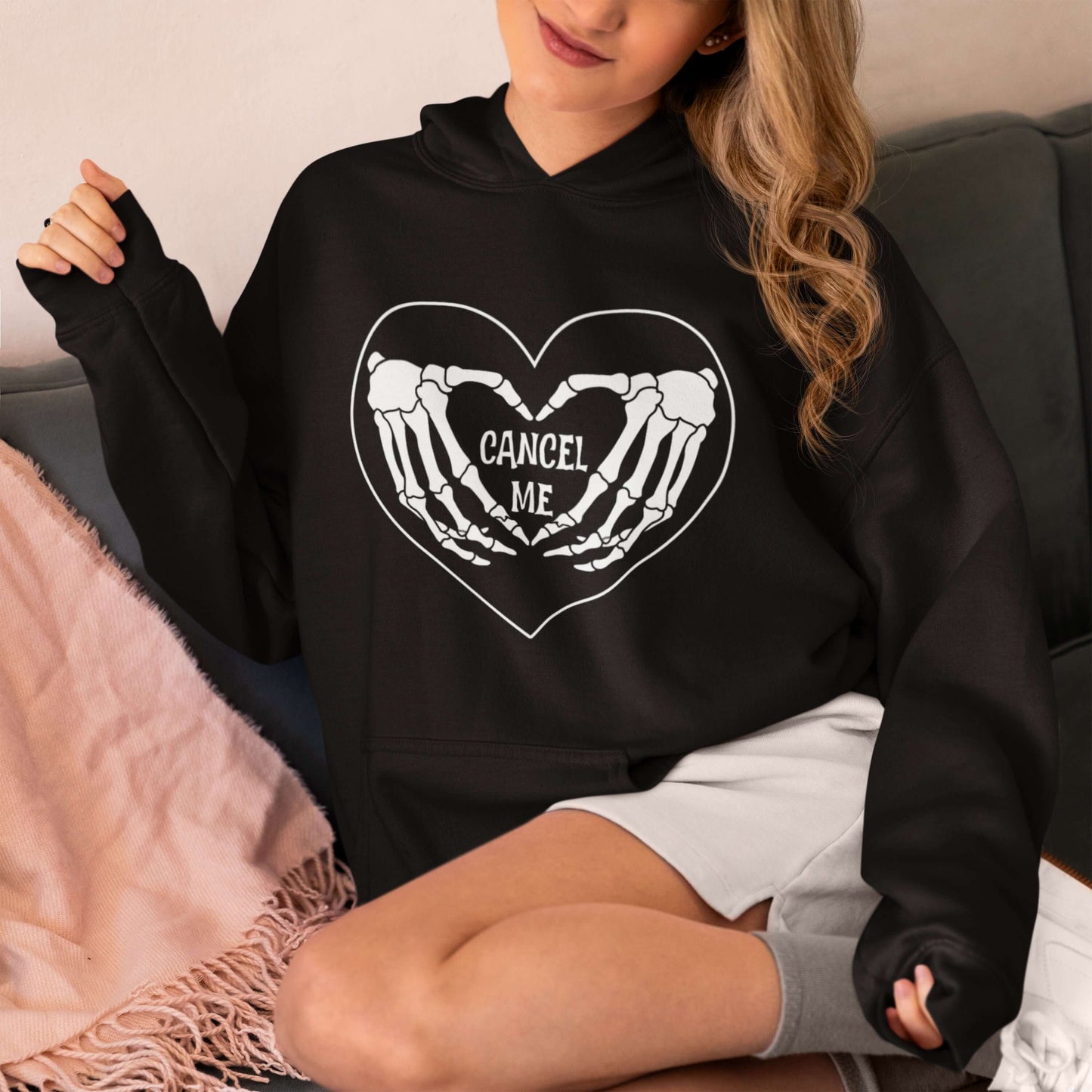 Woman wearing a black hoodie sweatshirt with a black heart and skeleton hands making a heart shape printed on the front. The words Cancel Me are inside of the heart.