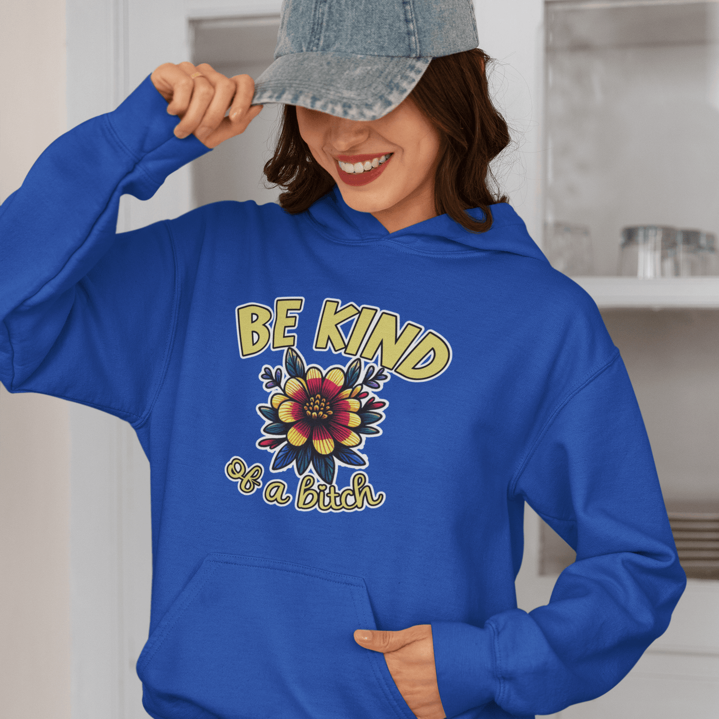 Woman wearing a bright blue hoodie sweatshirt with an image of a flower and the words Be kind above the flower in yellow bold block font. The words Of a bitch are smaller in script font under the flower. The design is printed on the front of the hoodie.