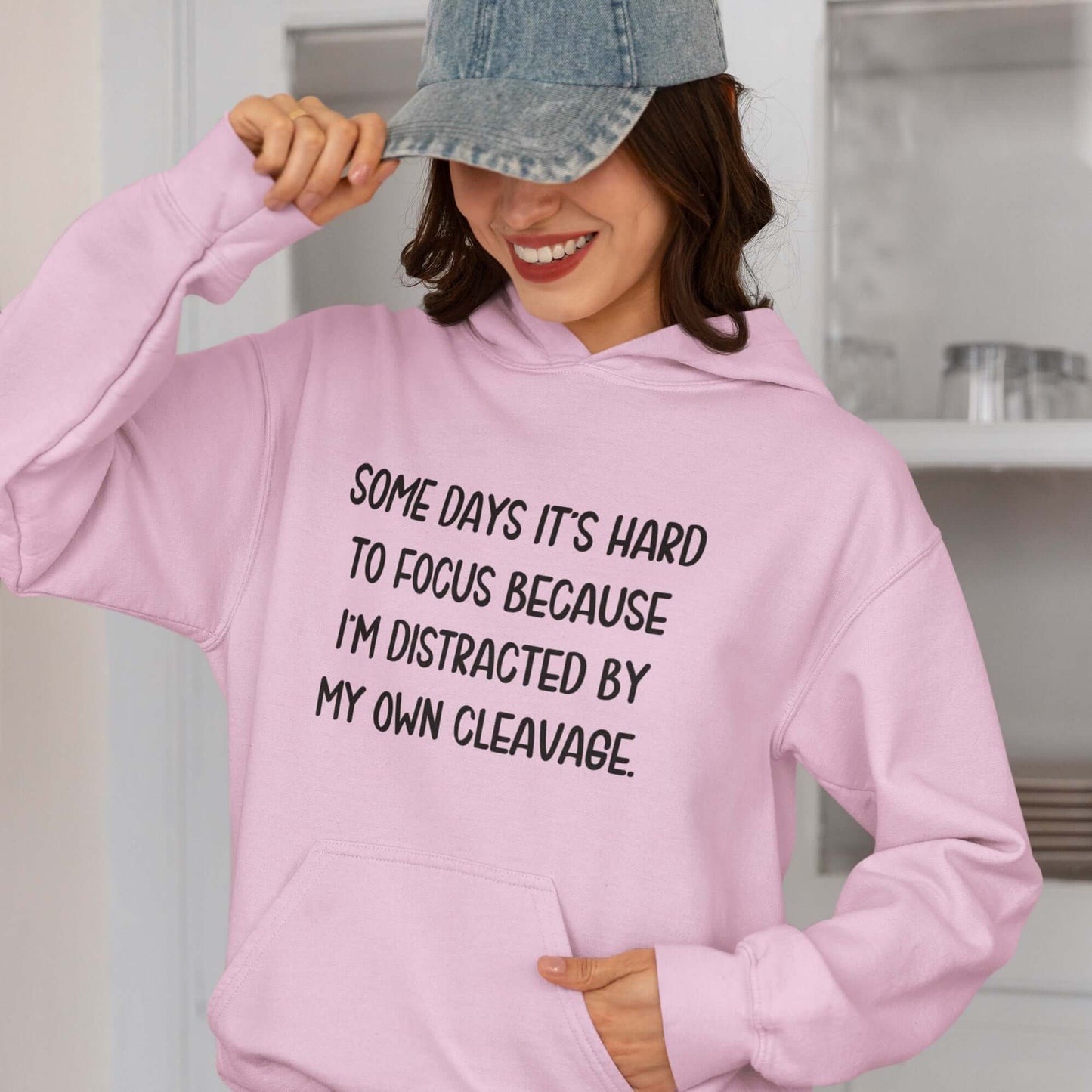 Woman wearing a light pink hoodie sweatshirt with the phrase Some days it's hard to focus because I'm distracted by my own cleavage printed on the front.