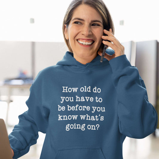 Woman wearing an indigo blue hoodie sweatshirt with the phrase How old do you have to be before you know whats going on question mark printed on the front.