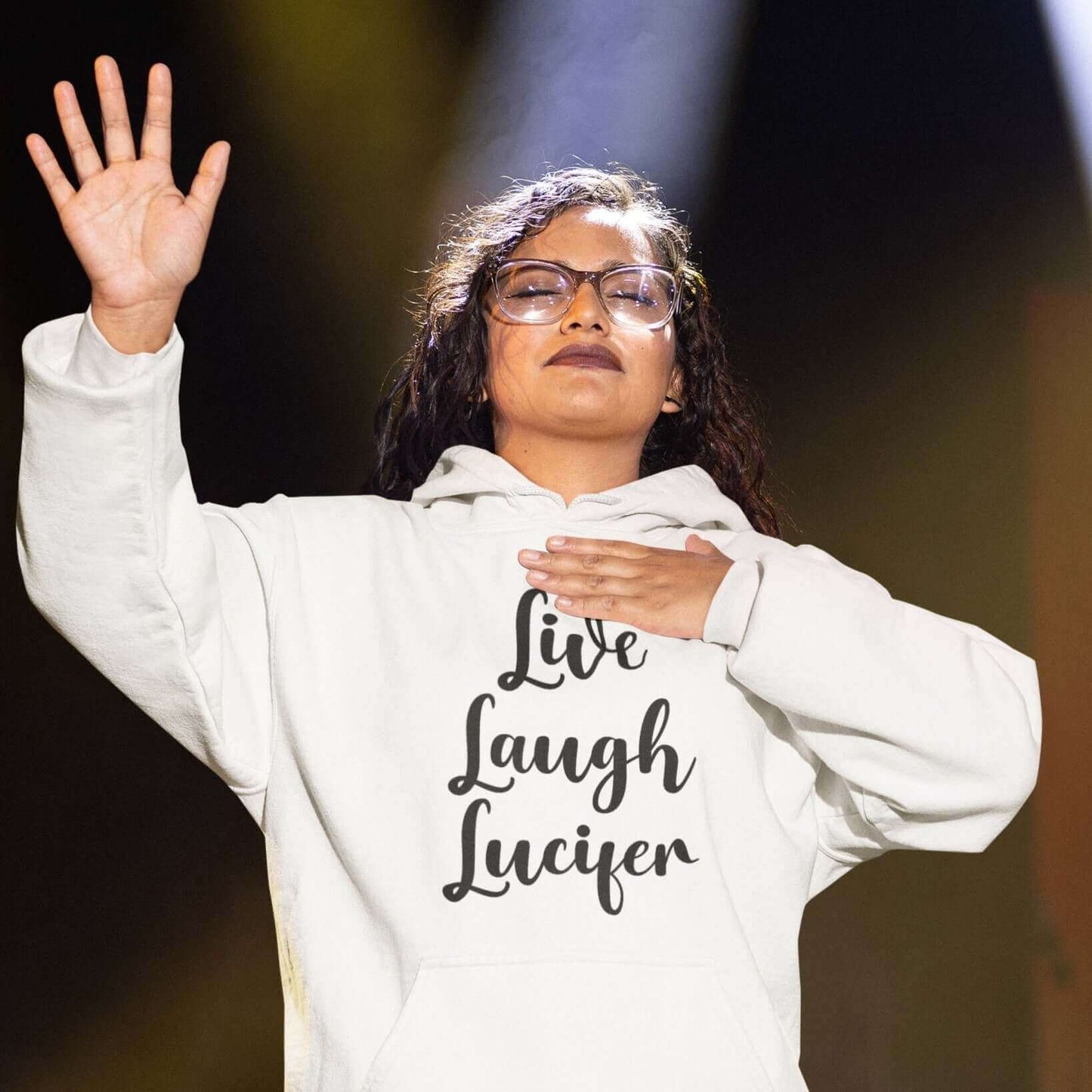 Woman praising while wearing a white hoodie sweatshirt with the parody phrase Live, Laugh, Lucifer printed on the front.