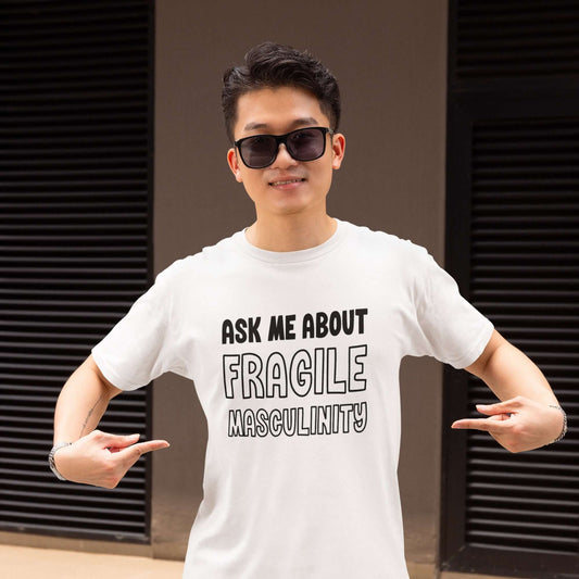 Man wearing sunglasses and a white t-shirt. He is pointing at the front of his shirt. The shirt has the phrase Ask me about fragile masculinity printed on the front.