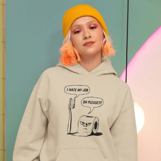 Woman wearing a sand hoodie with a cartoon graphic on the front. The cartoon is a line drawing of a toothbrush and roll of toilet paper. Both have speech bubbles above . The toothbrush says I hate my job and the toilet paper says Oh please.