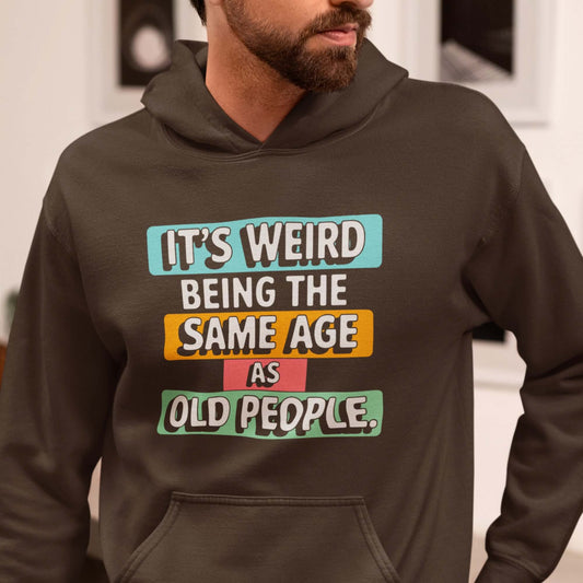 Man wearing a dark brown hoodie sweatshirt with colorful graphics that say It's weird being the same age as old people printed on the front.