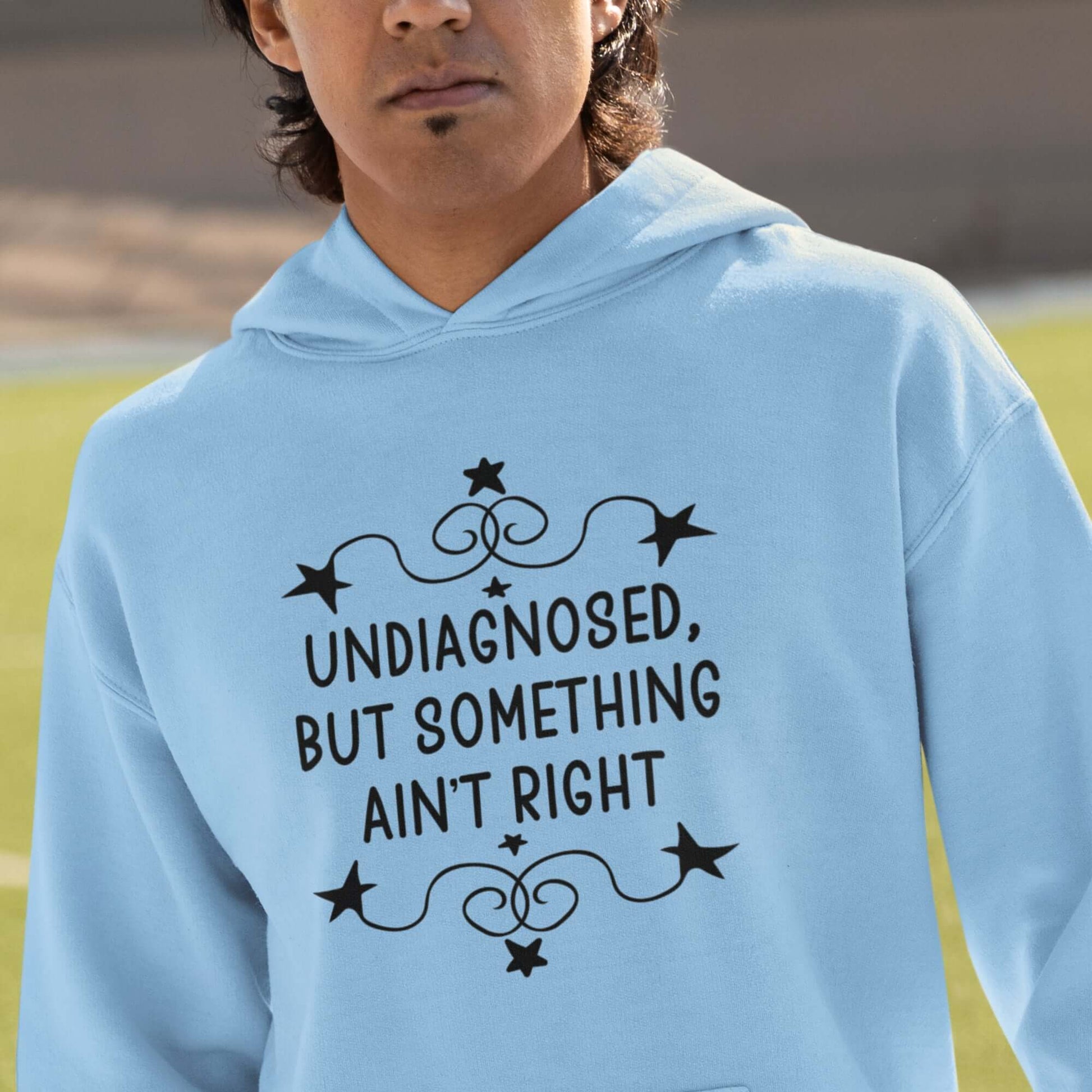 Man wearing a light blue hoodie sweatshirt with the phrase Undiagnosed, but something ain't right printed on the front. The text is black and has a star graphic embellishment around the words.