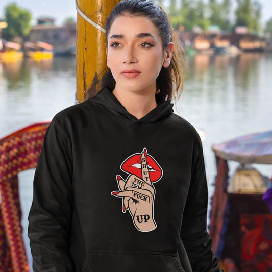 Woman wearing a black hoodie sweatshirt with an image of a hand over lips making the shh gesture. The words Shut the fuck up are printed on the hand. The graphics are printed on the front of the hoodie.