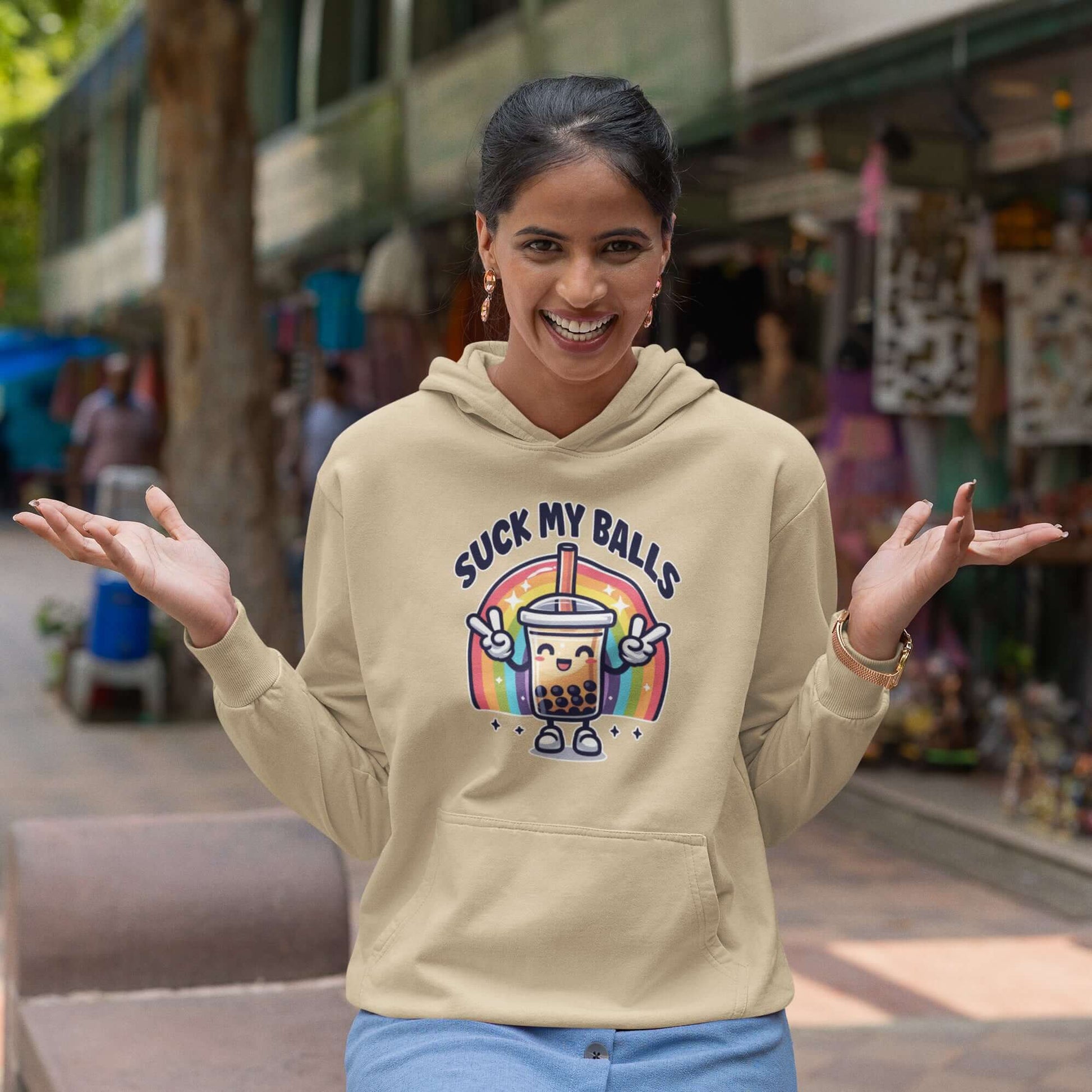 Woman wearing a sand color hoodie with graphics of a rainbow and a smiling boba bubble tea. The bubble tea has arms and legs. The phrase Suck my balls is printed above the rainbow. The graphics are on the front of the hoodie.