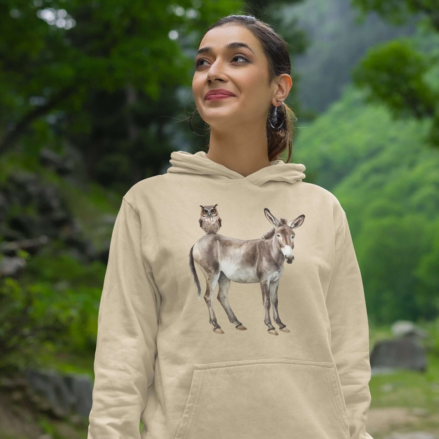 Smiling woman wearing a sand colored hoodie sweatshirt with image of a donkey with wise owl sitting on it. The graphic is printed on the front of the hoodie.