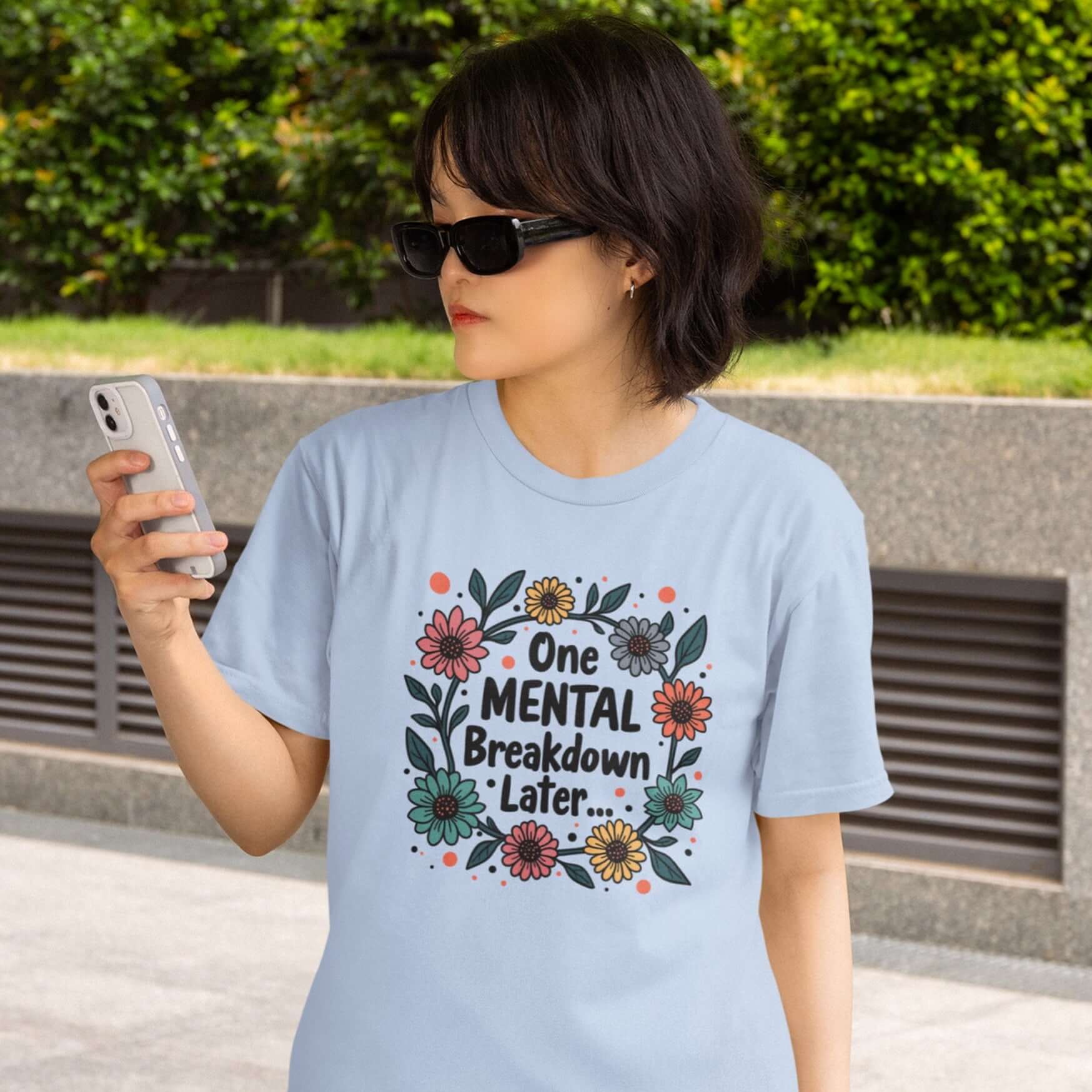 Woman wearing a light blue t-shirt with a multi color floral wreath design printed on the front. The words One mental breakdown later are printed in the center of the wreath. 