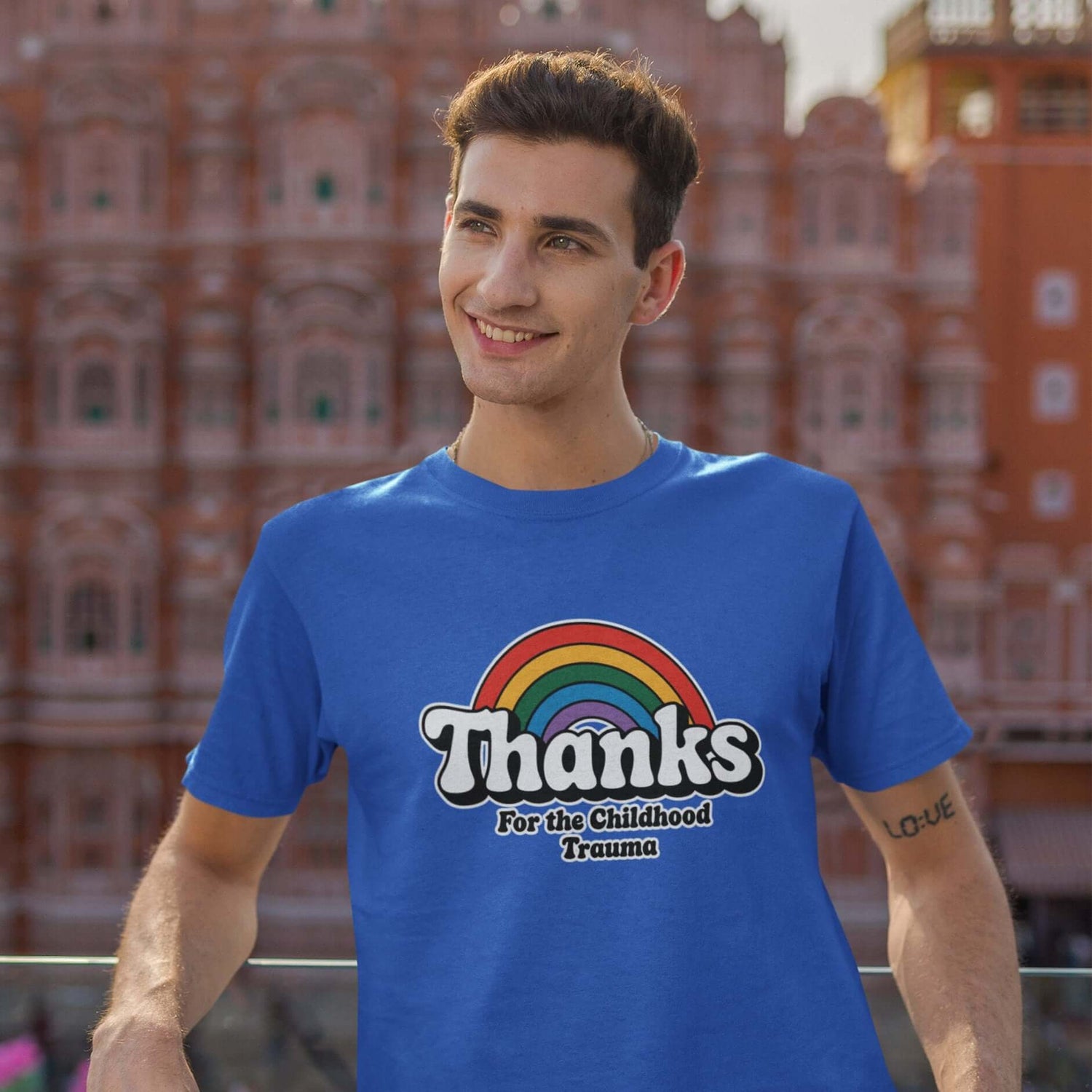 Man wearing a royal blue t-shirt with an image of a rainbow and the words Thanks for the childhood trauma printed on the front. The word Thanks is large and the rest of the words are smaller under.