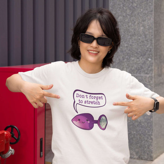 Woman wearing sunglasses and a white t-shirt. She is pointing to the graphics on the shirt. There is an image of a purple anal plug with a smile face with a speech bubble above with the words Don't forget to stretch. The graphics are printed on the front 