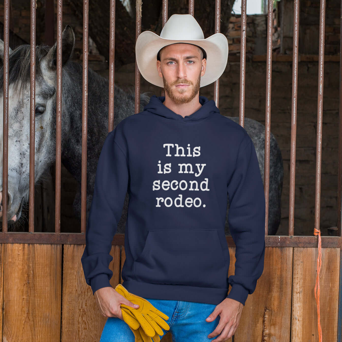 Man wearing a cowboy hat and a navy blue hoodie in a barn setting. There is a horse in the background. The hoodie sweatshirt has the funny phrase This is my second rodeo printed on the front.