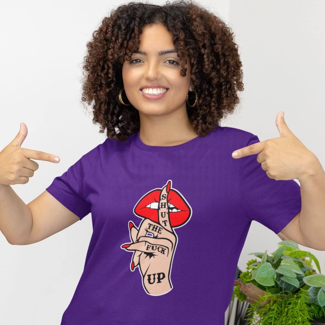 Woman wearing a purple t-shirt with an image of a hand over lips making the shh gesture. The words Shut the fuck up are printed on the hand. The graphics are printed on the front of the shirt.