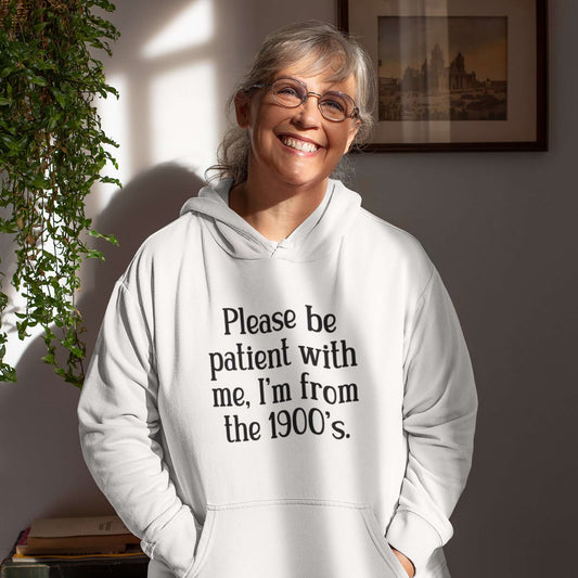 Middle aged woman wearing a white hoodie sweatshirt with the phrase Please be patient with me, I'm from the 1900s printed on the front.