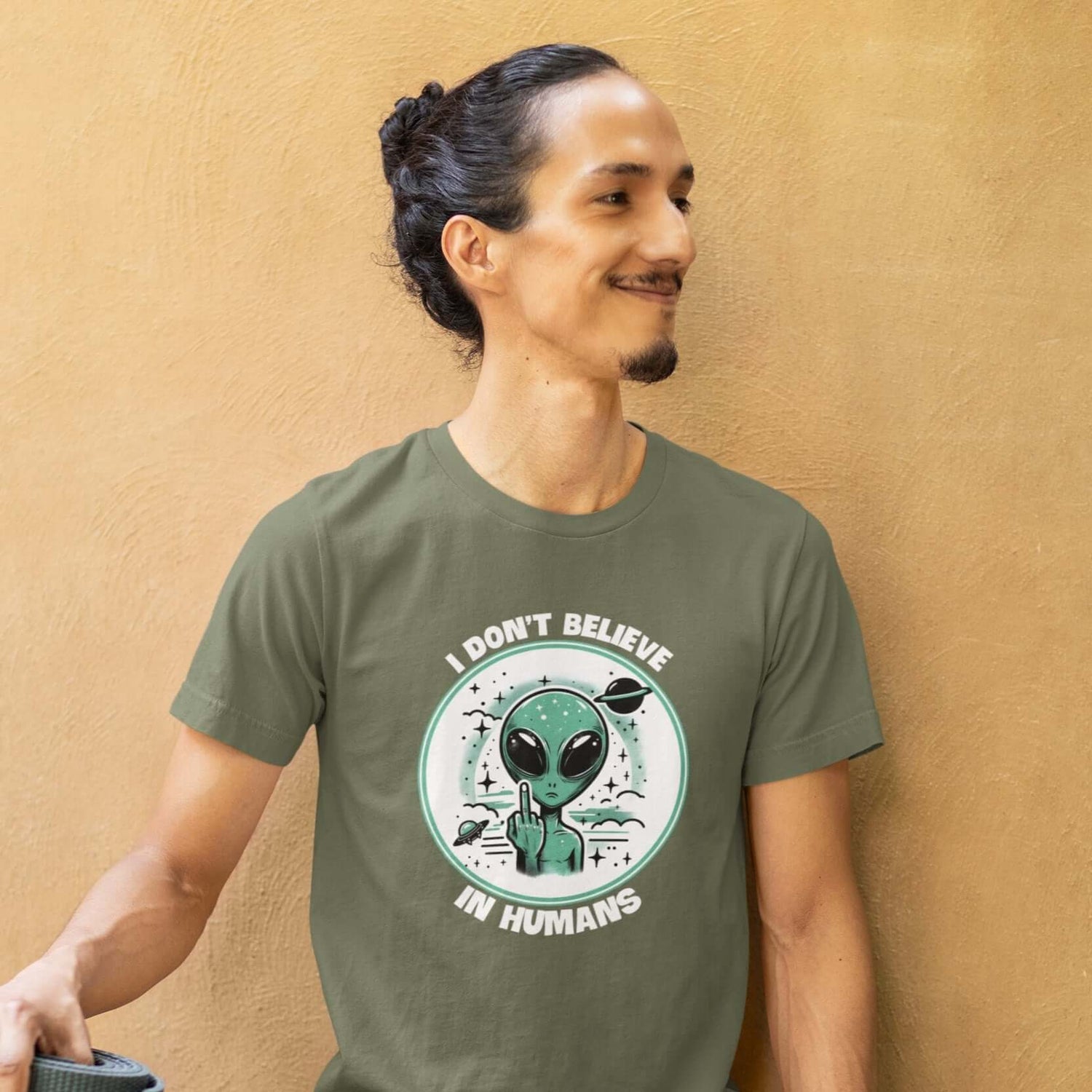 Man wearing a military green t-shirt with an image of an alien flipping the middle finger. The words I don't believe in humans are printed around the image. The graphics are printed on the front of the shirt.