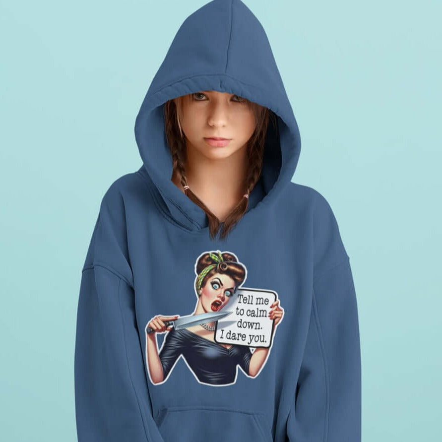 Woman wearing indigo blue hooded sweatshirt with the hood on. The sweatshirt has a graphic of an angry looking retro woman holding a knife and a sign. The sign says Tell me to calm down I dare you. The graphic is printed on the front of the hoodie.