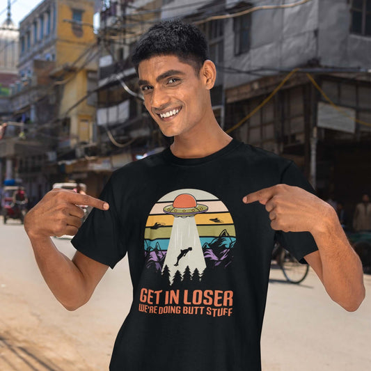 Man pointing at the funny black t-shirt he is wearing with UFO alien abduction graphic and the words Get in loser, we're doing butt stuff printed on the front.