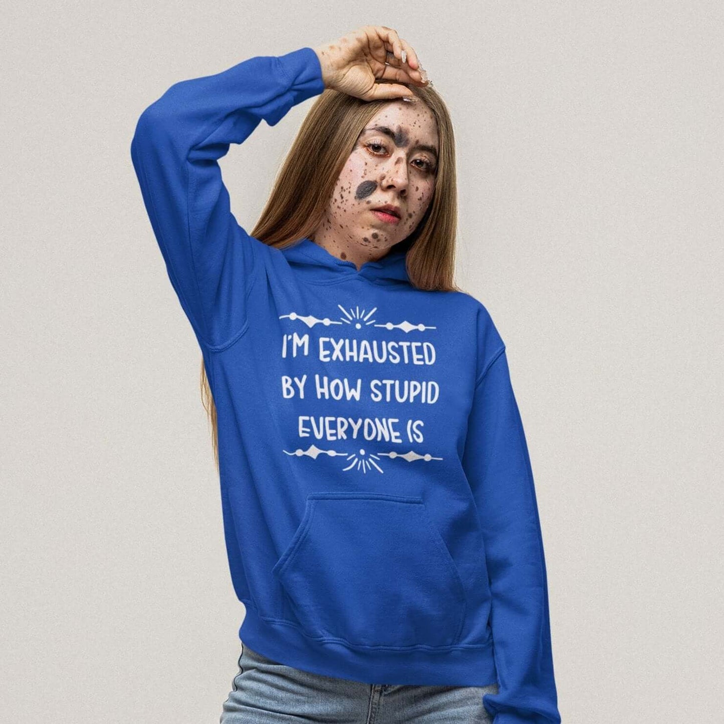 Woman wearing a royal blue hoodie sweatshirt with the phrase I'm exhausted by how stupid everyone is. The graphics are printed on the front of the hoodie.