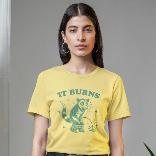 Woman wearing a yellow t-shirt with an image of an opossum peeing. The words It burns are printed above.