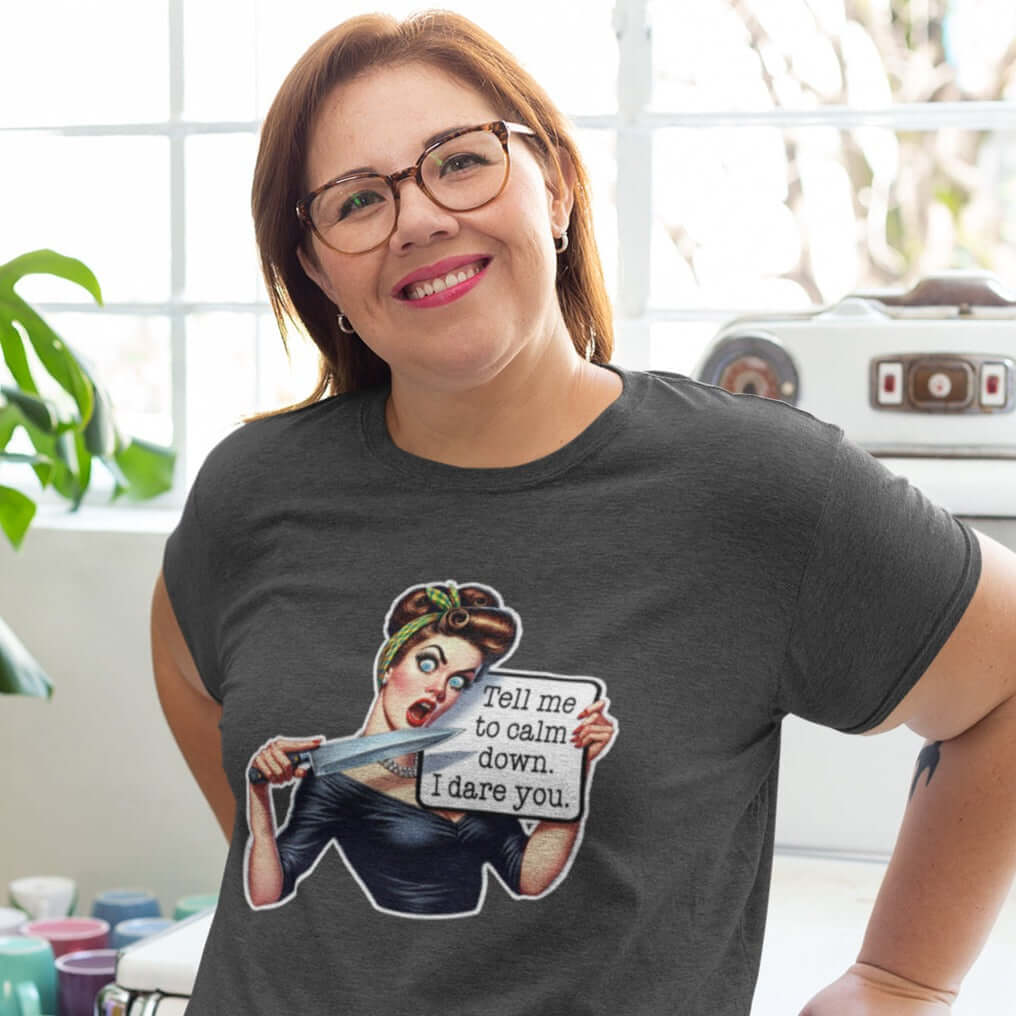 Woman wearing dark heather grey t-shirt. The t-shirt has a graphic of an angry looking retro woman holding a knife and a sign. The sign says Tell me to calm down I dare you. The graphic is printed on the front of the shirt.