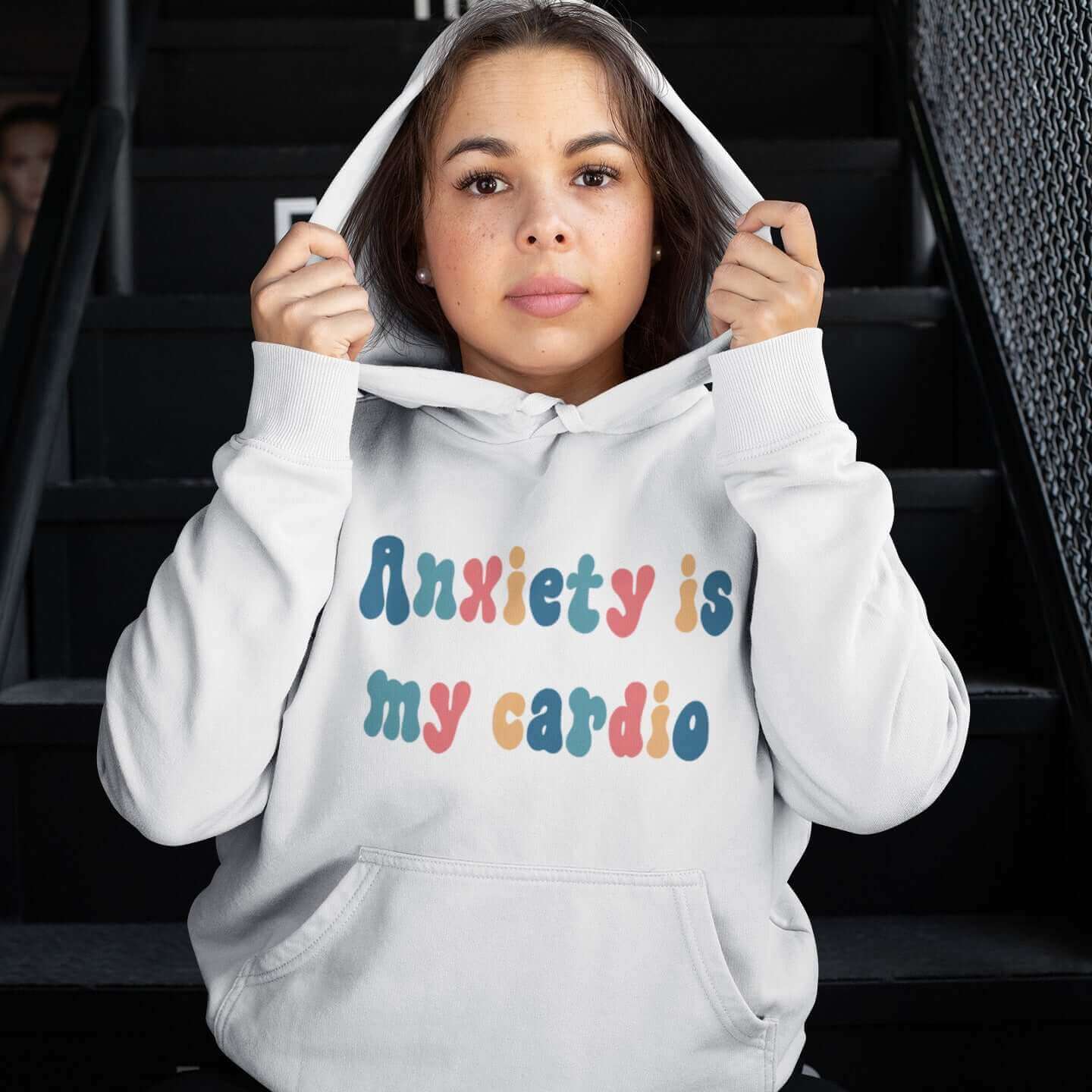 Woman wearing a white hoodie sweatshirt with the phrase Anxiety is my cardio printed in multiple colors on the front of the hoodie.