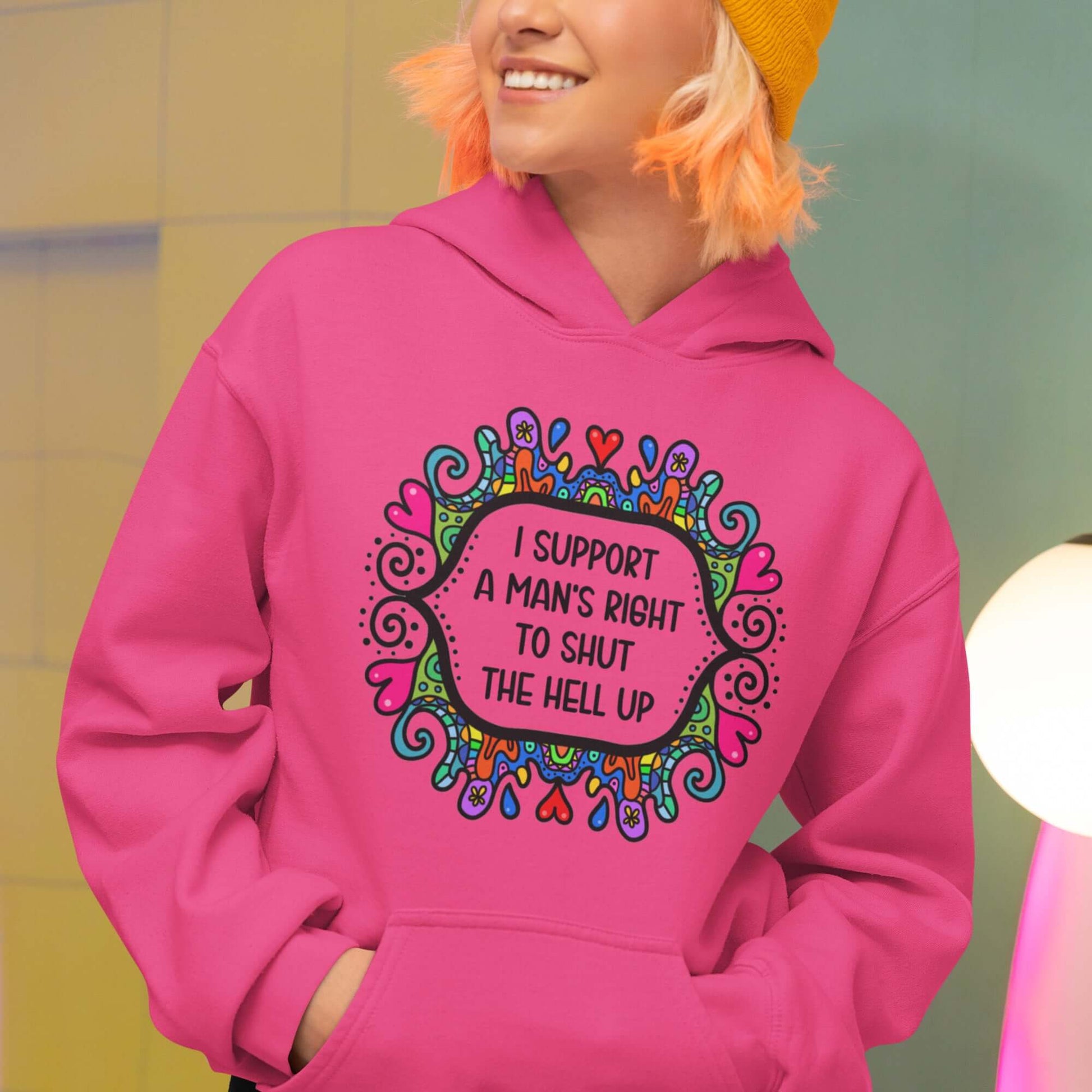 Woman wearing a heliconia pink color hoodie sweatshirt with a colorful doodle graphic and the words I support a man's right to shut the hell up printed on the front.