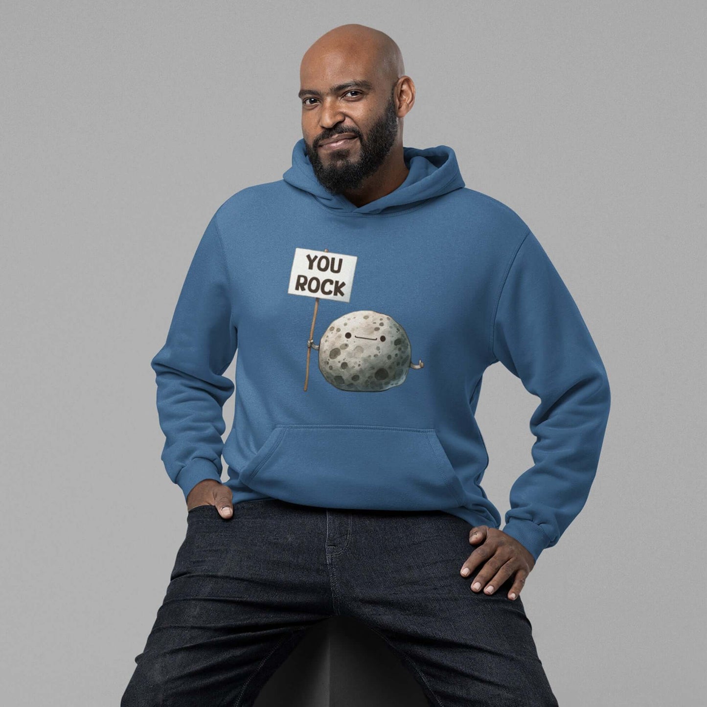 Man sitting on a stool wearing an indigo blue hoodie sweatshirt. The hoodie has an image of a grey rock that is holding a sign. The sign says You Rock. The image is printed on the front of the hoodie.