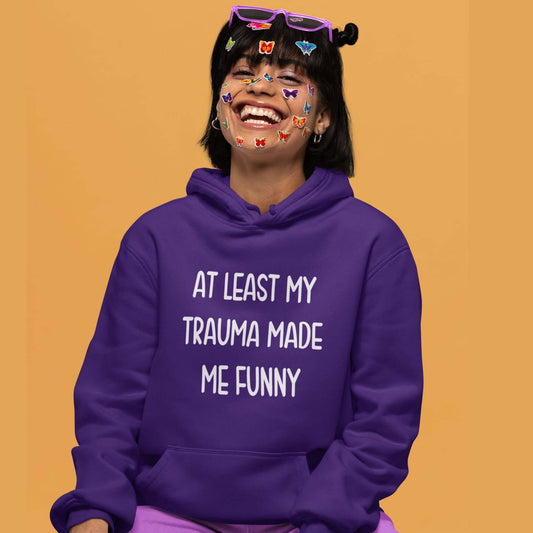 Woman wearing a purple hoodie sweatshirt with the words At least my trauma made me funny printed on the front.