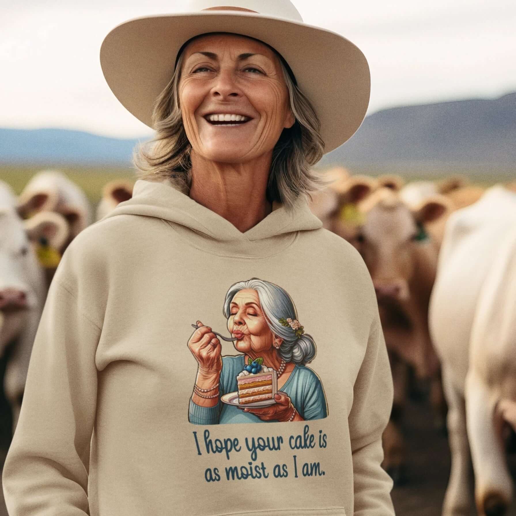 Older woman wearing sand colored hoodie sweatshirt with image of grey haired older woman enjoying a bite of a piece of cake with the words I hope your cake is as moist as I am printed on the front.