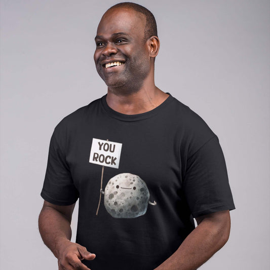 Smiling man wearing a black t-shirt that has an image of a grey rock that is holding a sign. The sign says You Rock. The image is printed on the front of the t-shirt..